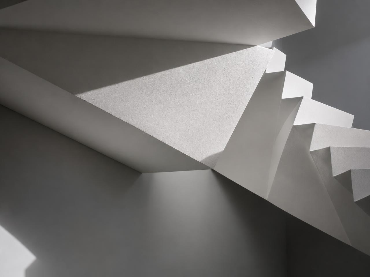 Folded white plaster soffit beneath a floating staircase in raking light